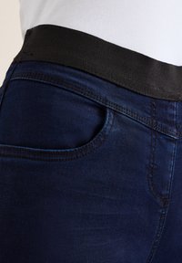 Donkerblauwe jeans met een gladde textuur, voorzien van een zwarte elastische tailleband en details van een voorzak, met nadruk op de stiksels en pasvorm.