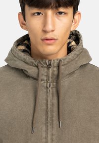 Veste à capuche zippée vert olive avec une surface texturée, des cordons de serrage et une doublure intérieure à carreaux. Design simple et fonctionnel.
