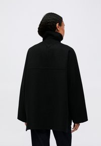 Person med kort, krøllet hår iført en løs, sort jakke med høj krave, står mod en ensartet lys baggrund og vender væk fra kameraet.