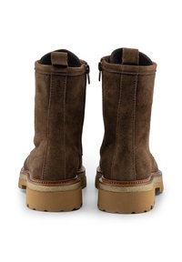 Par de botas de ante marrón hasta el tobillo con tiradores, cremalleras laterales y suelas de goma beige gruesas, vistas desde atrás sobre un fondo blanco.