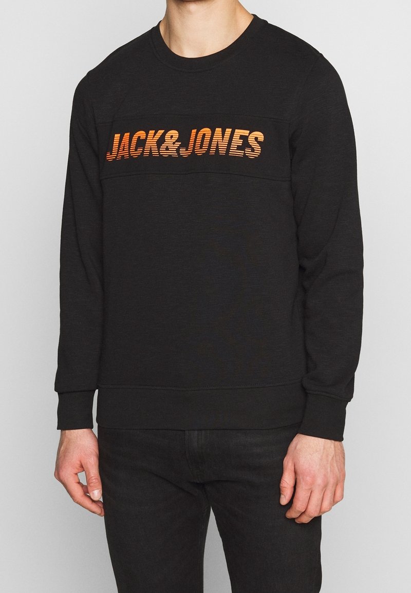Homme portant un sweat-shirt à manches longues noir avec le texte orange "JACK & JONES" sur la poitrine, associé à un pantalon noir sur un fond uni.