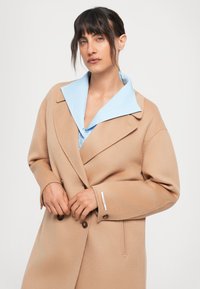 Donna con i capelli scuri che indossa un cappotto beige doppiopetto sopra una camicia azzurra a collare, su uno sfondo bianco uniforme.