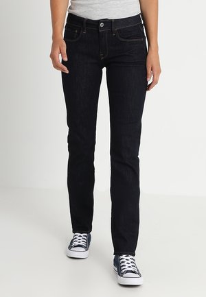 Personne portant un jean noir slim et des baskets noir et blanc à lacets, debout devant un fond blanc uni.