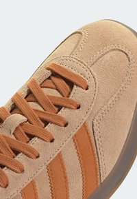 Beige mockasinsneaker med bruna snören och tre bruna ränder. Har zigzagmönster i sömmen på sidan och en gummisula.