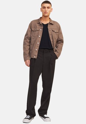 Mann trägt eine braune Jacke mit Knöpfen über einem schwarzen Hemd, lockere schwarze Hose und schwarz-weiße Turnschuhe, steht mit einer Hand in der Tasche.