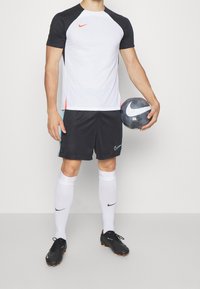 Ropa deportiva de Nike que incluye una camiseta blanca y negra con detalles rojos, pantalones cortos negros, calcetas blancas y botas de fútbol negras, sosteniendo un balón de fútbol.