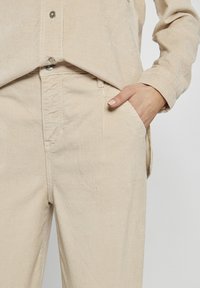 Beżowa koszula z korduroju z zapięciem na guziki, z kieszonką z przodu i teksturowanym materiałem. Połączona z spodniami z korduroju w tym samym kolorze, które mają kieszenie boczne.