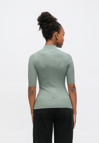 Ανοιχτό πράσινο ριμπ πλεκτό πουλόβερ με ψηλό γιακά και κοντά μανίκια, εφαρμοστή γραμμή, φορεμένο με μαύρο παντελόνι, που δείχνει υφή υφάσματος.