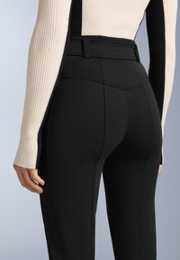 Fekete, testhez álló nadrág, textúrázott derékrésszel, sima anyaggal és függőleges varrásokkal. Ribbelt, krémszínű, hosszú ujjú felsővel viselve.