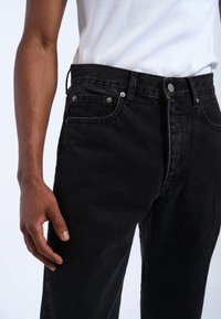 Jeans de denim negro con corte recto, diseño de cinco bolsillos, herrajes plateados y una textura sutil; combinados con una camiseta sin mangas de canalé blanca.