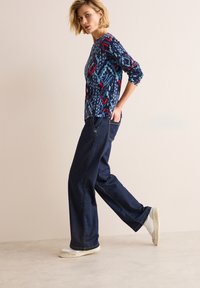 Langemouwtop met een geometrisch patroon in blauw, rood en wit; gecombineerd met losse, donkerblauwe denim broeken en witte sneakers.