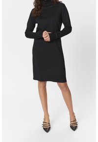 Schwarzes Rollkragenkleid mit langen Ärmeln und gerippter Struktur. Knielang, figurnah geschnitten. Kombiniert mit schwarzen spitzen Pumps mit dekorativen Riemen.