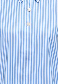 Tissu vertical rayé bleu et blanc avec deux boutons décoratifs, l'un semblable à une perle et l'autre carré et doré, près d'un col plié.