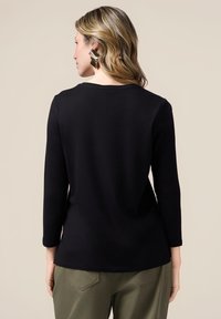Maglia nera a maniche lunghe con scollatura rotonda, texture liscia e vestibilità rilassata, abbinata a pantaloni verde oliva e grandi orecchini geometrici.