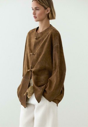 Femme aux cheveux blonds courts portant une veste en daim marron oversize avec des fermetures à bascule et un pantalon blanc ample sur un fond uni.