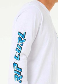 Langærmet hvid T-shirt med blå "RIP CURL" grafik på venstre ærme. Har rund halsudskæring og blød stoftekstur.