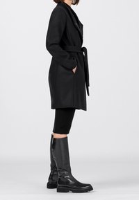 Musta villasekoitustakki, jossa on pitkät hihat, vyötäröllä solmittava nauha ja taskut, yhdistetty polvipitkiin mustiin nahkasaappaisiin, joissa on kuvioidut pohjat.