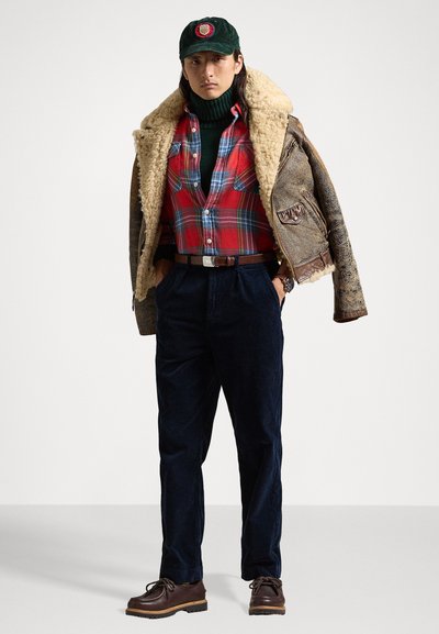 Brun shearling-fodrad jacka, röd och blå rutig skjorta, mörkgrön polokrage, mörka kordflöjlsbyxor och bruna snörskor. Grön keps med logotyp.