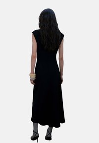Rochie neagră fără mâneci cu un corset ajustat și tiv fluid, realizată dintr-un material neted. Accesorizată cu o brățară mare de culoare crem.