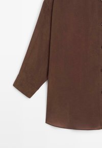 Camicia marrone a maniche lunghe realizzata in tessuto morbido. Presenta una chiusura con bottoni, polsini ampi e un orlo leggermente curvato.