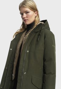 Parka vert olive avec fermeture éclair à l'avant, capuche ajustable par cordon de serrage et doublure intérieure matelassée. Dotée de boutons-pression et de deux poches latérales. Tissu texturé.