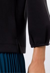Top nero con maniche a tre quarti a sbuffo, realizzato in un tessuto liscio, con orlo dritto e spacchi laterali. Abbinato a una gonna plissettata color teal.