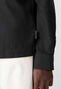 Tela de chaqueta negra con una textura suave, que presenta una etiqueta cerca del dobladillo. Primer plano de la muñeca que revela la costura y una camisa blanca contrastante debajo.