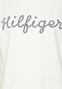 Bílý bavlněný tričko s černým provázkovým písmem, které nápis "Hilfiger" napříč přední částí. Měkká textura s jednoduchým designem.