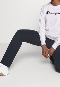 Person som bär en vit Champion-tröja, marinblå byxor och vita sneakers, knästående med händerna vilande på låret mot en plain bakgrund.