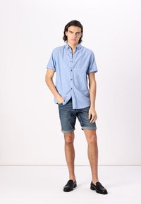 Chemise boutonnière à manches courtes bleu clair, shorts en jean et mocassins noirs. La chemise présente un tissu texturé avec une coupe décontractée.