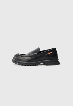 Mocasines de cuero negro con punta redondeada, tacón bajo y suela de goma gruesa y texturizada. Presentan un detalle de costura y un pequeño logo en el lateral.