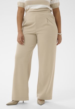 Femme portant un pantalon beige à jambes larges, un haut beige à rayures, des talons pointus beiges et un bracelet argenté au poignet droit.