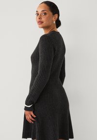 Frau mit Sommersprossen, die ein dunkelgraues geripptes Strickkleid trägt, silberne Ohrringe und ein silbernes Armband, und über ihre Schulter schaut vor einem schlichten Hintergrund.