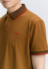 Camisa polo marrón con un cuello texturizado a rayas en tonos rojo y marrón oscuro. Logo bordado con "1962" en el pecho.