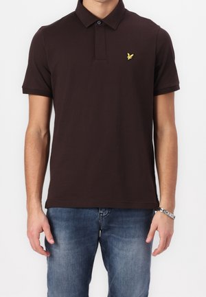 Mand iført en mørkebrun kortærmet poloshirt med en gul fuglelogo på venstre bryst og blå jeans.