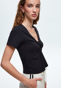 Top nero a maniche corte con scollo a V e dettaglio arricciato sul petto, dotato di un accento dorato. Tessuto liscio, design corto.