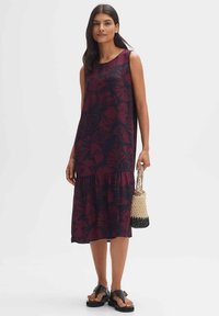 Ärmelloses, knielanges Kleid in Dunkellila mit floralem Muster. Ausgestellter Saum mit gestuftem Design. Model trägt eine geflochtene Tasche.