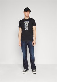Svart t-shirt med grafiskt tryck, som kombineras med mörkblå denimjeans och svarta sneakers. Modellen bär också en svart keps.