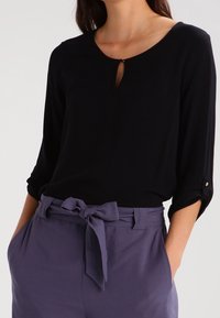 Svart blus med rund halsringning och nyckelhålsdetalj, i kombination med lätta, lila högmidjade byxor och ett knutet midjebälte.