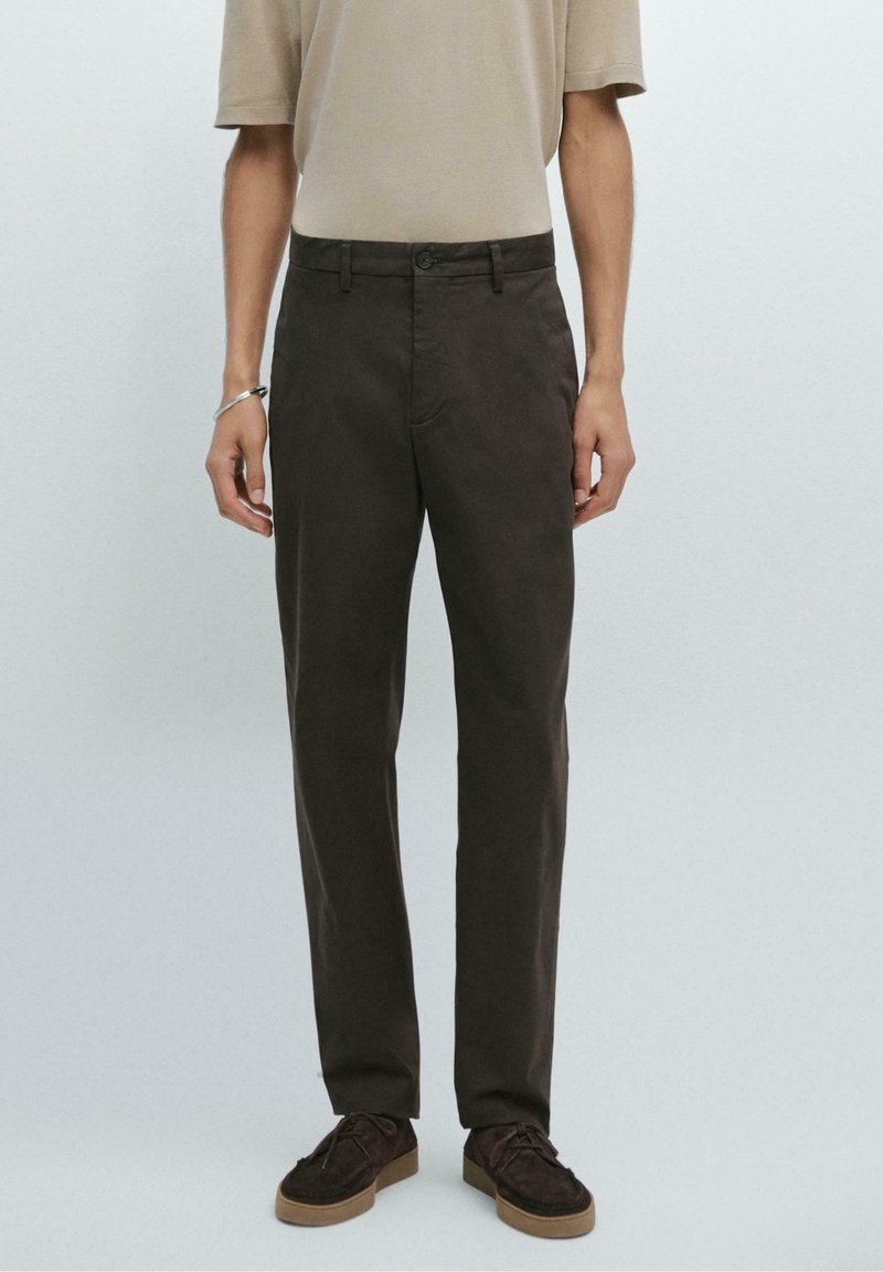 Homme portant un pantalon droit marron foncé, une chemise beige à manches courtes, des chaussures en daim marron et un bracelet en argent, debout devant un fond uni.