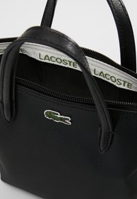 Czarna torebka na zamek błyskawiczny wykonana z teksturowanego materiału, z dwiema rączkami i wyraźnym logo Lacoste z przodu. Wnętrze ma markowe wykończenie.