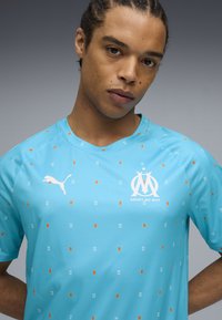 Gaiši zils futbola krekls, kas izgatavots no sintētiskā materiāla, ar oranžiem un baltiem ģeometriskiem rakstiem, Puma logo un izteiktu komandas emblemu.