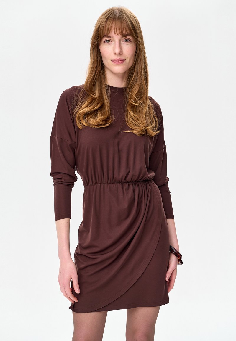 Młoda kobieta o długich, jasnobrązowych włosach, ubraną w brązową sukienkę kopertową z długim rękawem i ciemną bransoletką na nadgarstku, na białym tle.