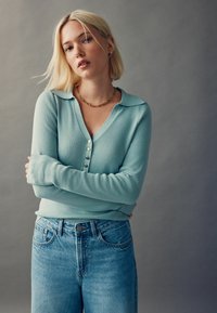 Pull en tricot côtelé bleu clair avec un col et une fermeture à boutons sur le devant, associé à un jean en denim bleu taille haute. Texture douce et coupe ajustée.
