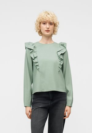 Kobieta z krótkimi kręconymi blond włosami ubrana w jasnozieloną bluzkę z długim rękawem ozdobioną falbanami z przodu oraz ciemnoszare jeansy, stoi na białym tle.