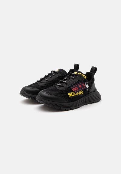 Paire de baskets noires sportives avec une tige en mesh, des semelles épaisses, des languettes et un lettrage jaune et rouge sur les côtés.