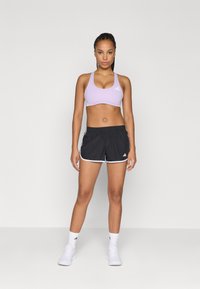 Sostenido deportivo de color púrpura claro con tirantes gruesos y acentos blancos, combinado con pantalones cortos negros con ribete blanco y el logo de tres rayas.