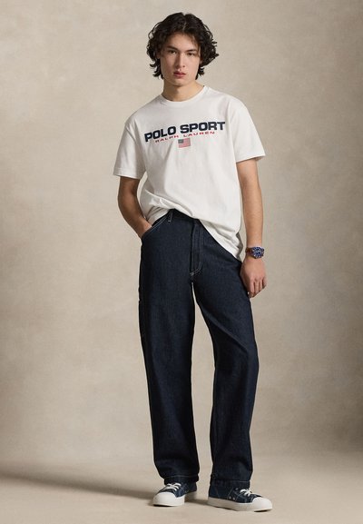 Άσπρο βαμβακερό μπλουζάκι με το γράφημα "POLO SPORT", μπλε τζιν παντελόνια, σκούρα μπλε αθλητικά παπούτσια και ένα ρολόι χειρός, στέκεται σε ουδέτερο φόντο.