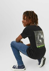 Svart t-shirt med ett grafiskt design av en jordglob och texten "EN BÄTTRE VÄRLD." Kombinerat med blå jeans och marinblå sneakers.