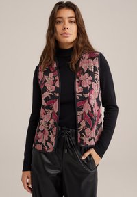 Cvjetni prsluk od tamnog materijala s ružičastim i crvenim botaničkim vezom; složen preko crne rolke, u kombinaciji s crnim, sjajnim hlačama.